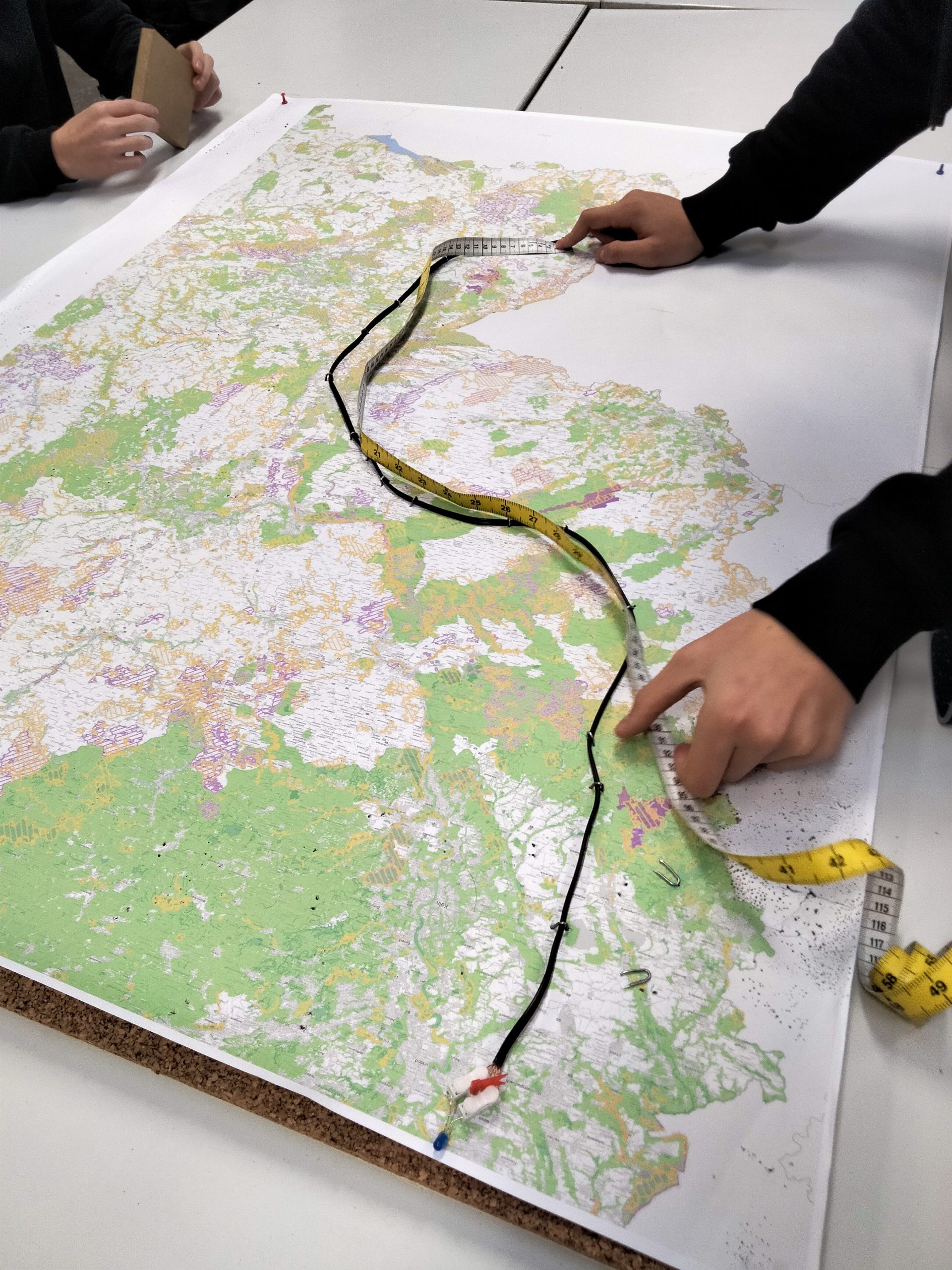 Hände zeigen auf eine detaillierte topografische Karte mit einem Maßband, das zur Messung von Entfernungen verwendet wird. Die Karte enthält verschiedene Farben, die unterschiedliche Geländearten darstellen.