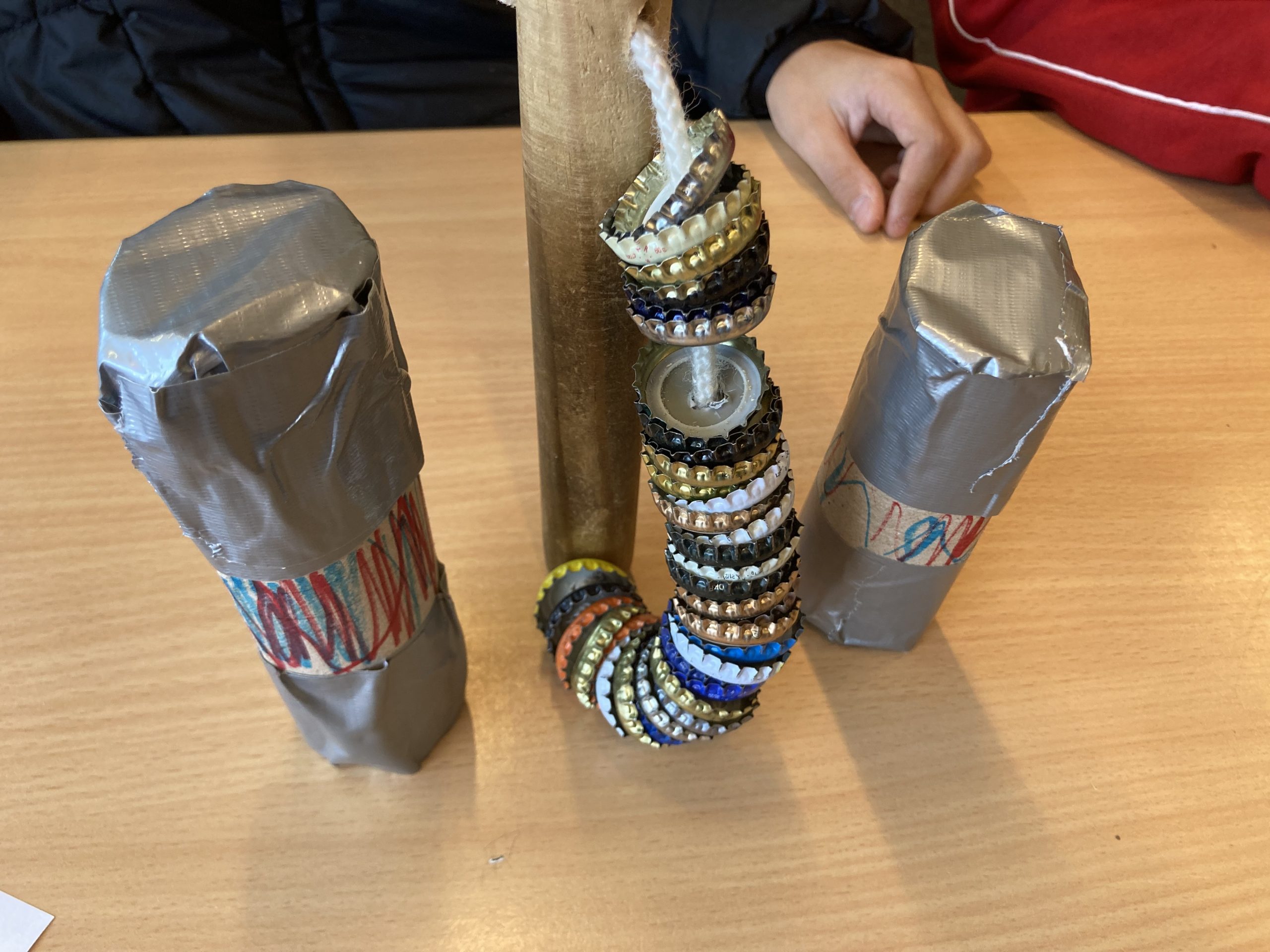 Ein handgefertigtes Spielzeug aus einem Holzstück und mehreren bunten Ringen, die auf einer Schnur aufgereiht sind. Zwei mit Silberfolie umwickelte Objekte stehen daneben, auf einem Tisch.