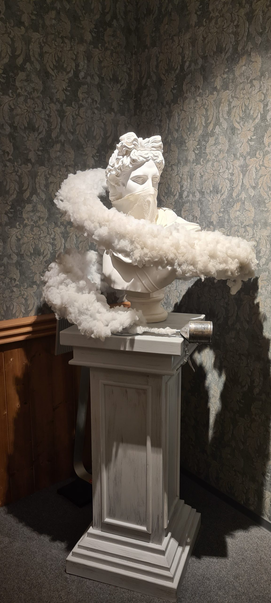 Moderne Skulptur eines Kopfes mit einer Wolkenformation aus weißen Federn, auf einem eleganten Sockel präsentiert, vor einer dekorativen Wand.