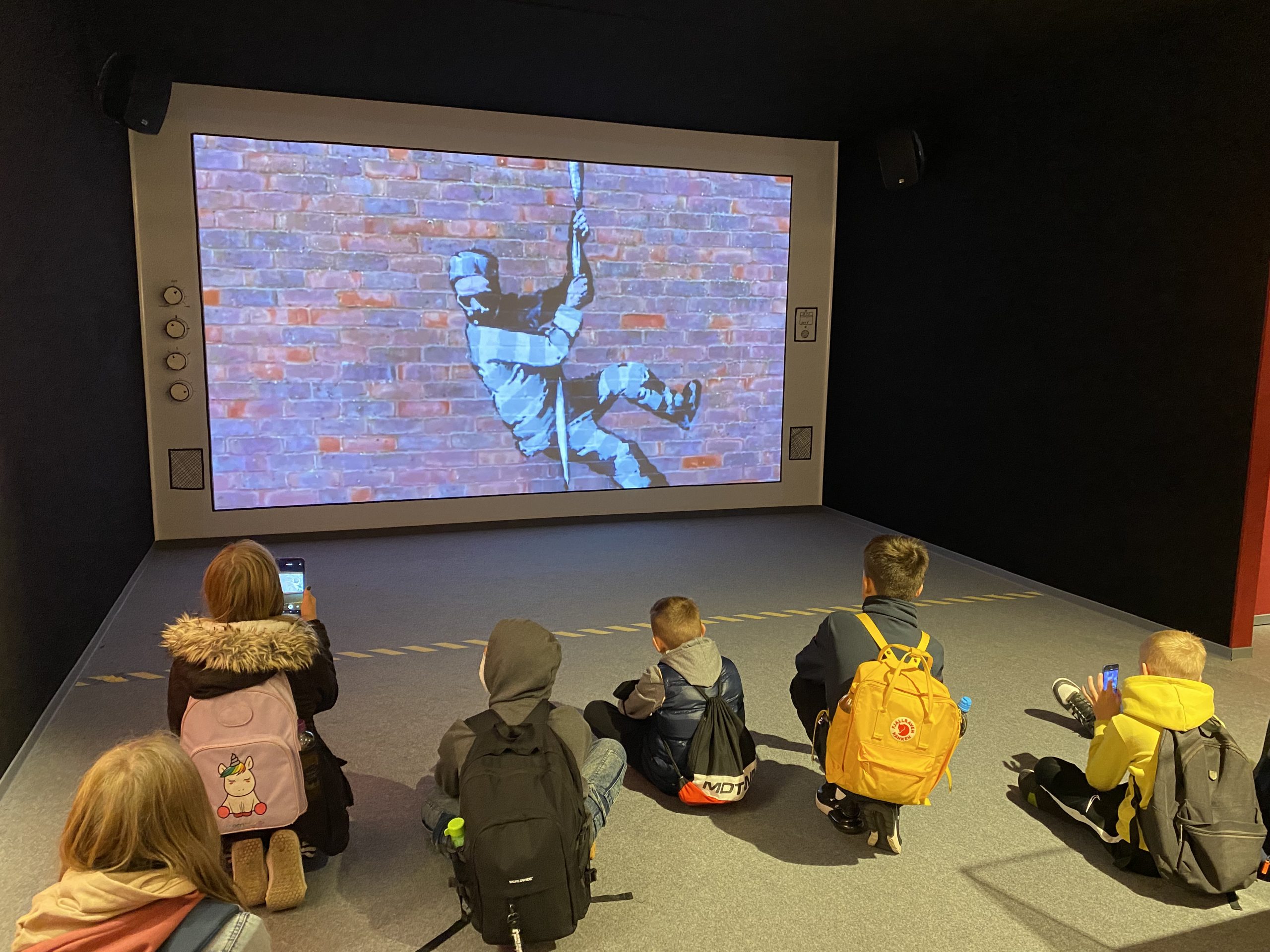 Eine Gruppe von Kindern sitzt auf dem Boden und schaut sich eine Projektion an, die einen Kletterer an einer Backsteinmauer zeigt. Die Szene vermittelt ein Gefühl von Abenteuer und Faszination für die dargestellte Aktivität.