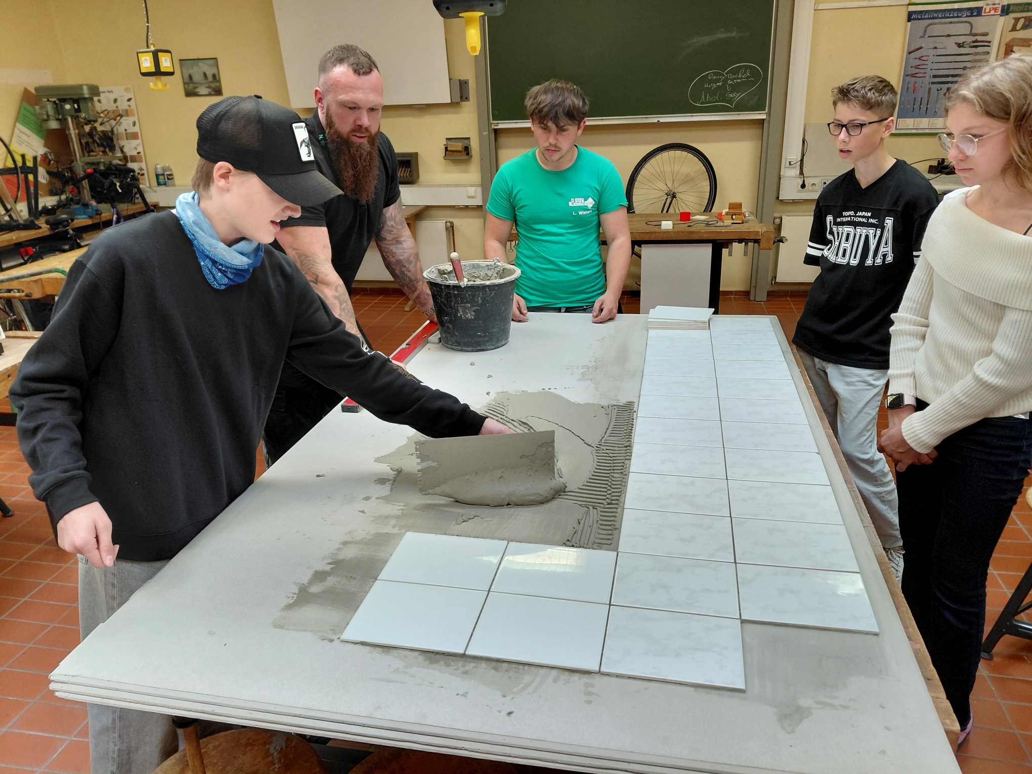 Schüler lernen das Fliesenlegen in einer Werkstatt, während ein Lehrer Anweisungen gibt. Auf dem Tisch liegen Fliesen und Mörtel, während die Gruppe aufmerksam zuschaut.