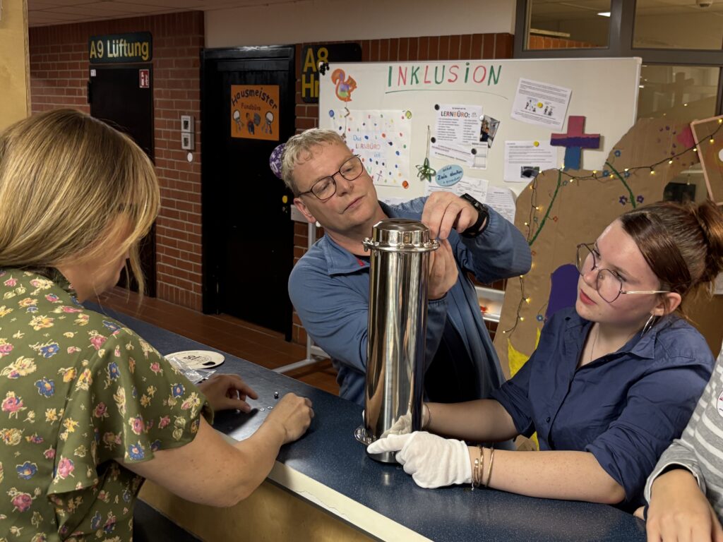 Drei Personen arbeiten zusammen an einem metallischen Objekt in einem Lernraum. Im Hintergrund sind Informationen zur Inklusion und Lernangeboten sichtbar. Die Szene zeigt Engagement und Teamarbeit in einer Bildungsumgebung.