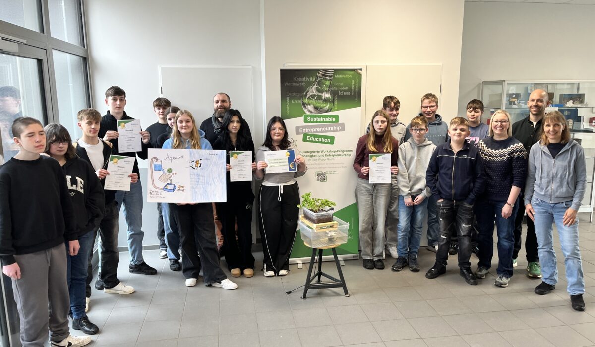 Gruppe von Schülern und Lehrern bei einer Zeremonie zur Verleihung von Zertifikaten für nachhaltige Unternehmensbildung. Die Teilnehmer halten stolz ihre Urkunden und stehen vor einem Plakat, das das Programm für nachhaltiges Unternehmertum bewirbt.