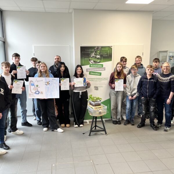 Gruppe von Schülern und Lehrern bei einer Zeremonie zur Verleihung von Zertifikaten für nachhaltige Unternehmensbildung. Die Teilnehmer halten stolz ihre Urkunden und stehen vor einem Plakat, das das Programm für nachhaltiges Unternehmertum bewirbt.