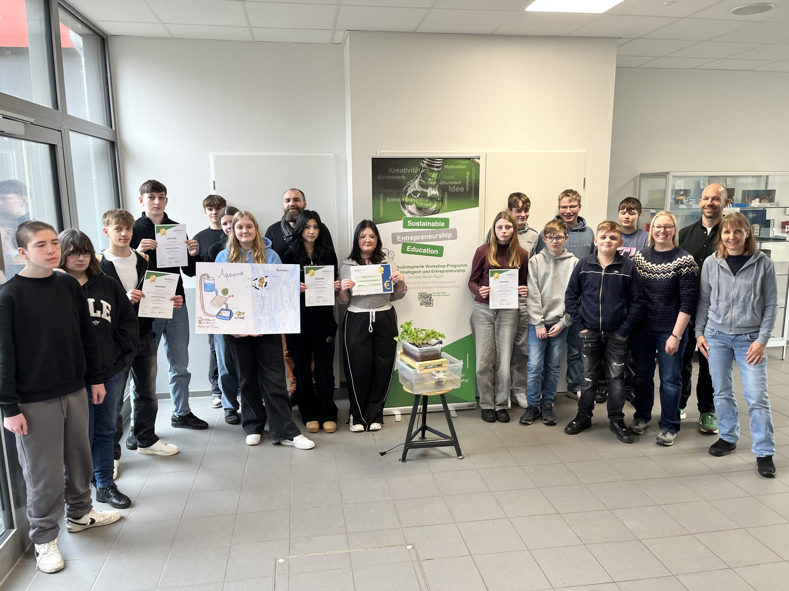Gruppe von Schülern und Lehrern bei einer Zeremonie zur Verleihung von Zertifikaten für nachhaltige Unternehmensbildung. Die Teilnehmer halten stolz ihre Urkunden und stehen vor einem Plakat, das das Programm für nachhaltiges Unternehmertum bewirbt.