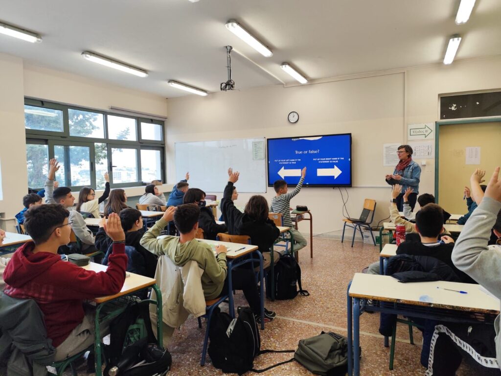 Schüler in einem Klassenzimmer heben die Hände zur Teilnahme an einer Diskussion, während eine Lehrerin an der Tafel steht und eine Präsentation über "Wahr oder Falsch" zeigt.