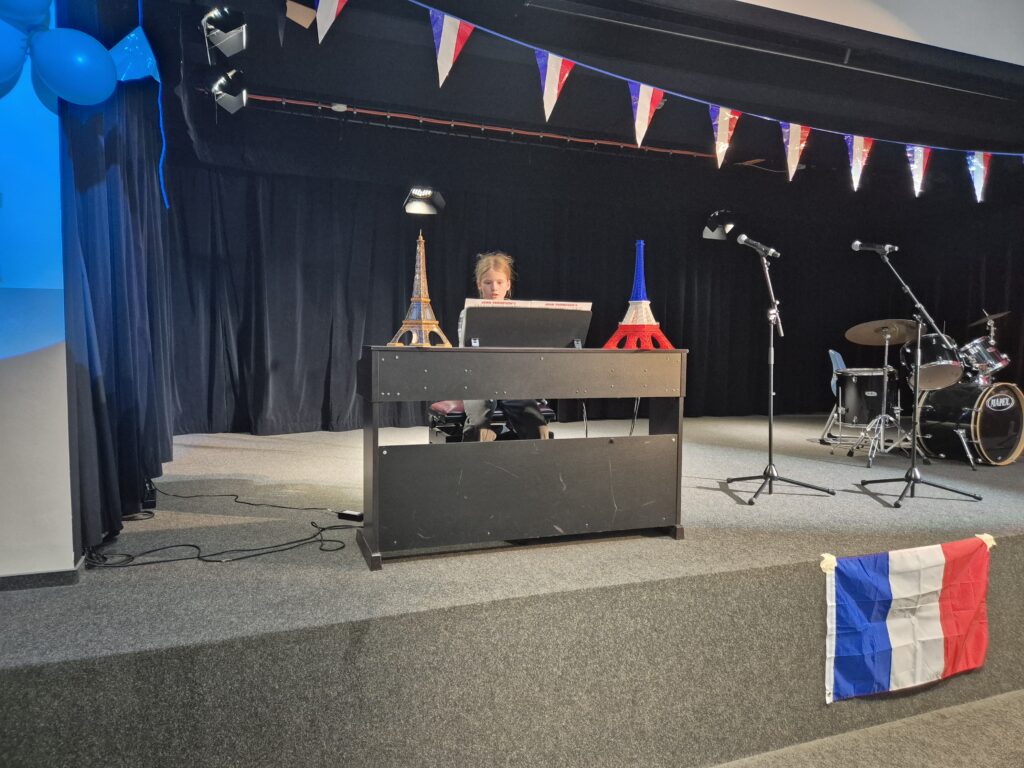 Ein Junge sitzt an einem Schreibtisch auf einer Bühne, umgeben von französischen Dekorationen, darunter Miniatur-Eiffeltürme und eine französische Flagge. Im Hintergrund sind Mikrofone und ein Schlagzeug sichtbar, was auf eine bevorstehende Aufführung hinweist.