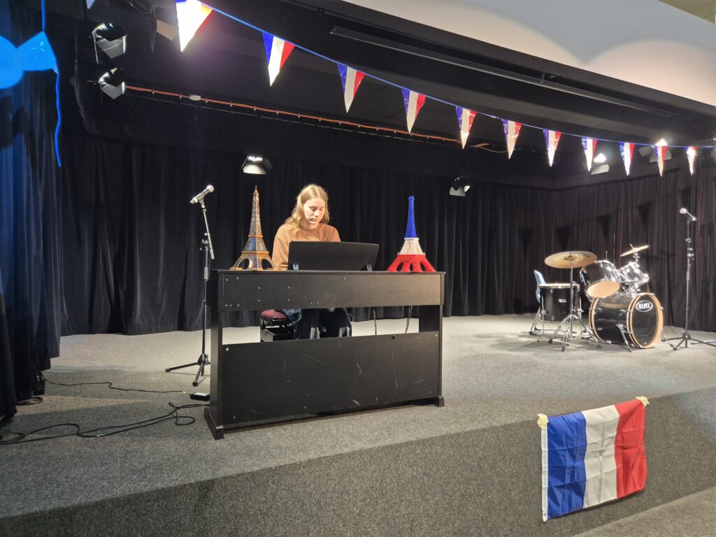 Ein junger Mann sitzt an einem Tisch auf einer Bühne, umgeben von französischem Dekor, darunter Miniatur-Eiffeltürme und eine französische Flagge. Im Hintergrund steht ein Schlagzeugset. Die Bühne ist mit bunten Wimpeln geschmückt und von Scheinwerfern beleuchtet, was auf eine bevorstehende Aufführung hinweist.