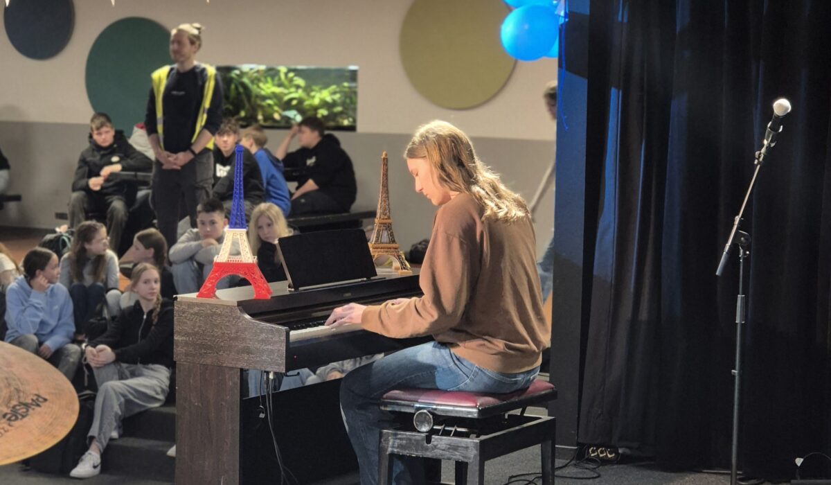 Schüler spielt Klavier bei einer Schulaufführung, umgeben von Mitschülern, mit Dekorationen in den Farben der französischen Flagge und kleinen Eiffeltürmen auf dem Klavier.