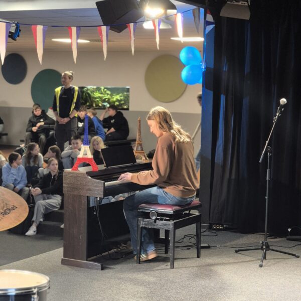 Schüler spielt Klavier bei einer Schulaufführung, umgeben von Mitschülern, mit Dekorationen in den Farben der französischen Flagge und kleinen Eiffeltürmen auf dem Klavier.