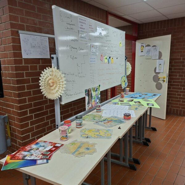 Tisch mit Kunstprojekten und Materialien in einem Klassenraum, darunter Zeichnungen, Farbkarten und Gläser mit Bastelmaterialien, vor einer Tafel mit Notizen und Informationen.