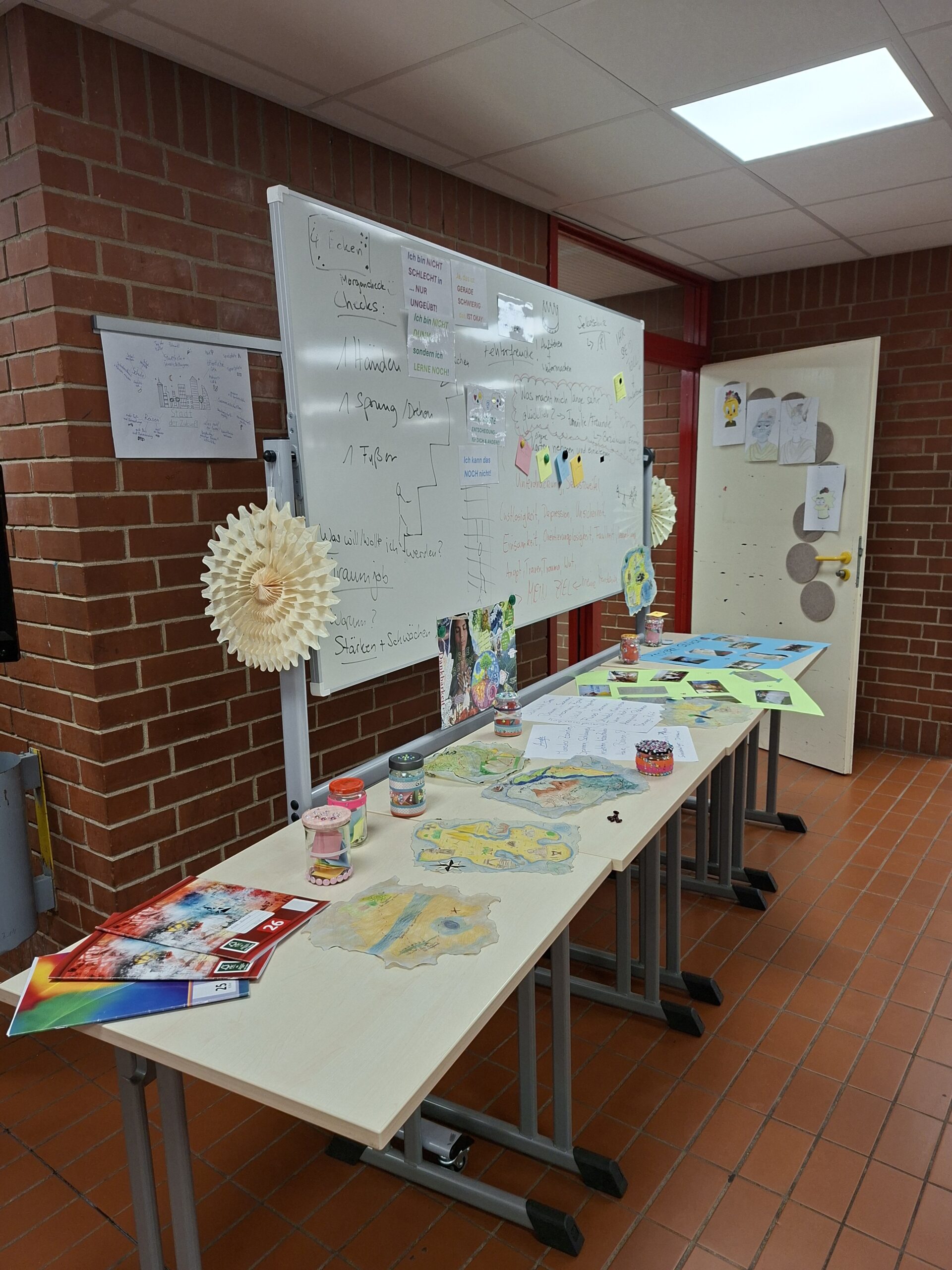 Tisch mit Kunstprojekten und Materialien in einem Klassenraum, darunter Zeichnungen, Farbkarten und Gläser mit Bastelmaterialien, vor einer Tafel mit Notizen und Informationen.