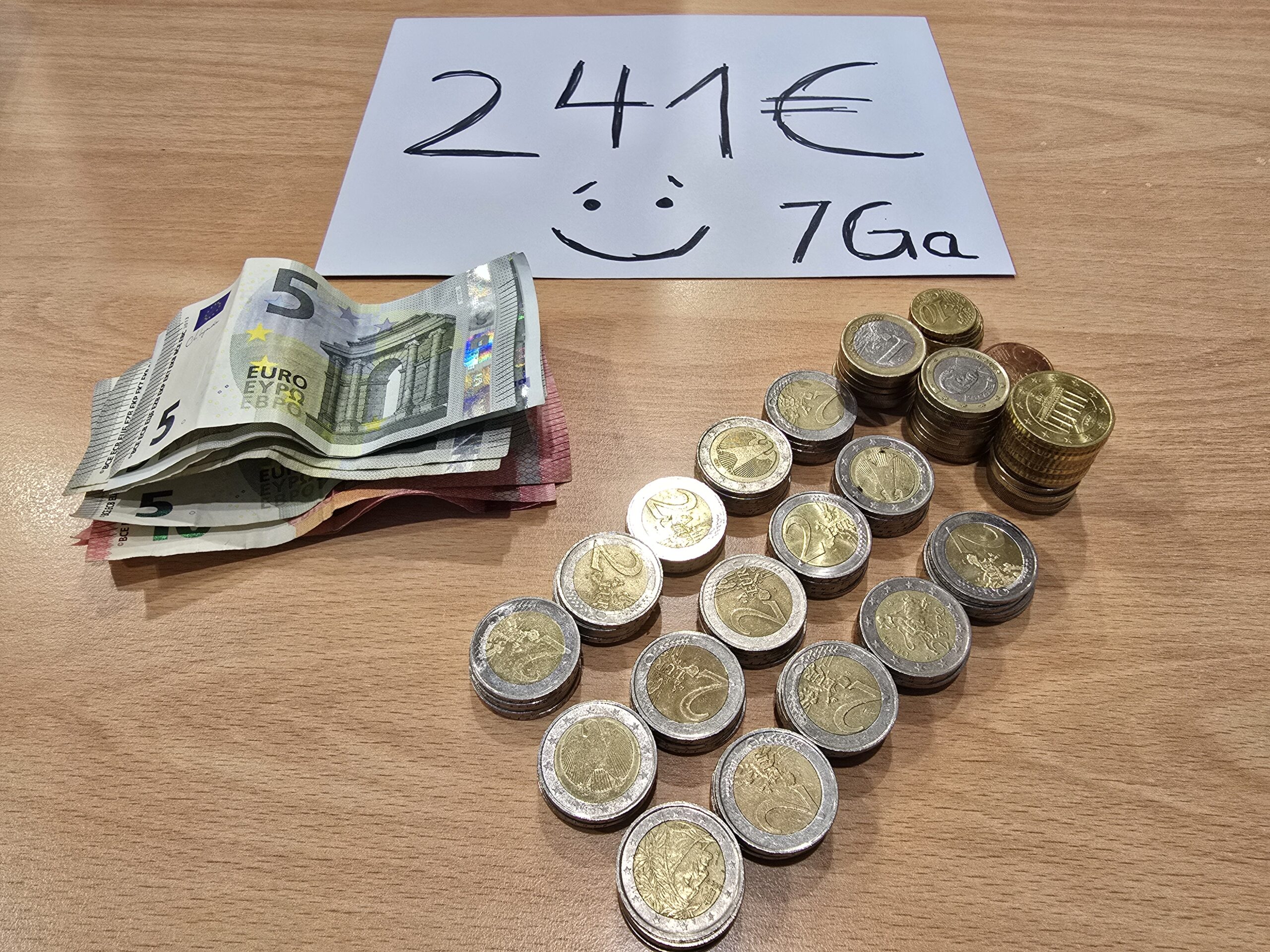 Stapel von Euro-Münzen und Scheinen auf einem Holztisch, mit einer Beschriftung von 241 € und einer Smileygrafik.