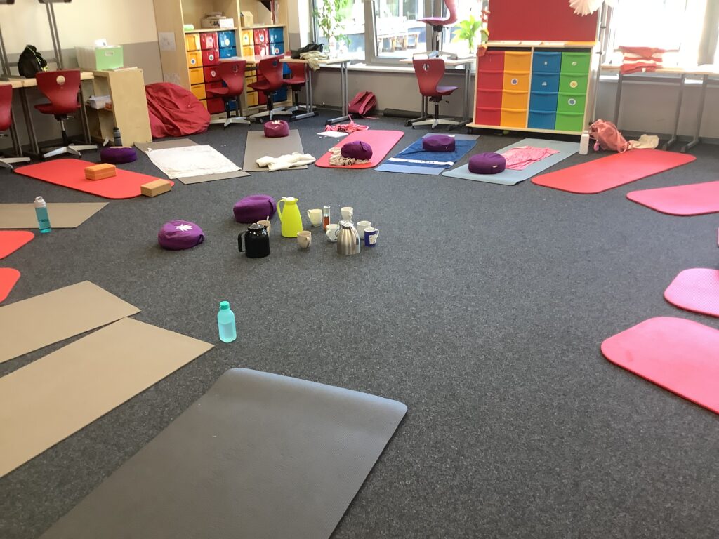 Yoga-Raum mit roten und grauen Matten, Meditationskissen, Wasserflaschen und Teekannen. Der Raum ist hell und einladend, ideal für Entspannung und Meditation.