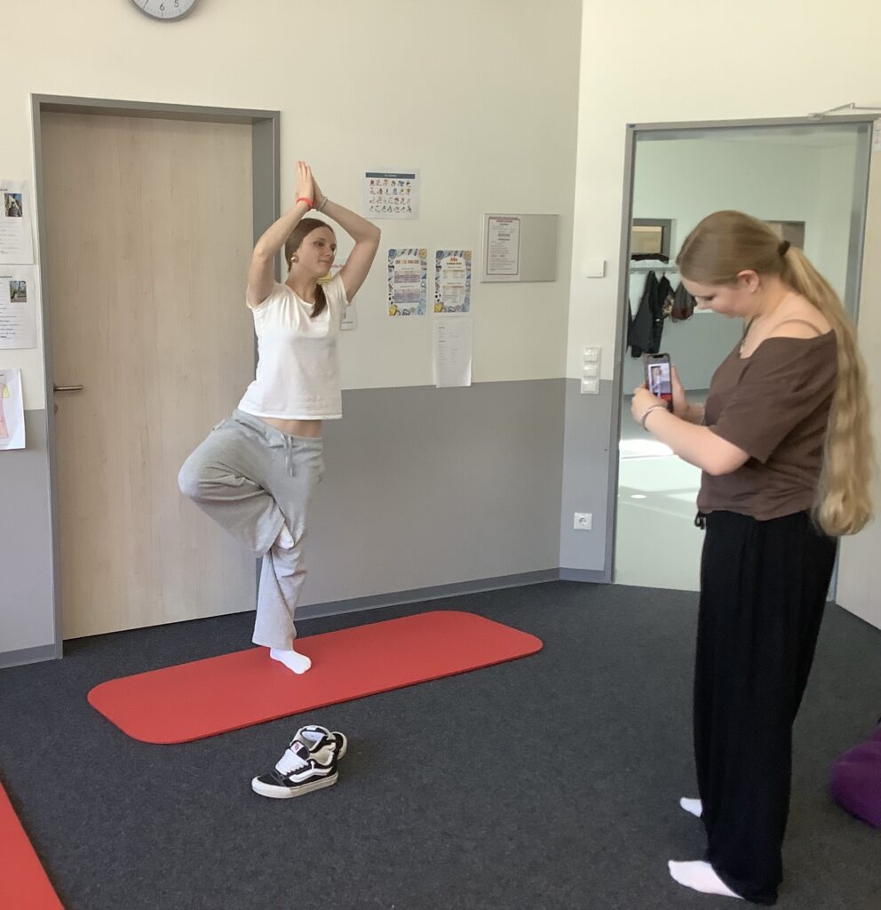 Zwei Frauen in einem Raum mit Sportmatten; eine Frau steht in einer Yoga-Pose auf einem Bein, während die andere sie mit einem Smartphone fotografiert. Im Hintergrund sind Wandtafeln und eine Tür sichtbar.