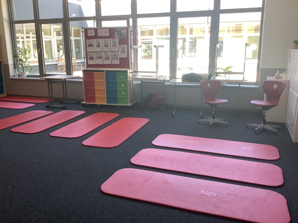 Ein heller Raum mit mehreren roten Yogamatten auf einem dunklen Teppichboden. Im Hintergrund steht ein bunter Schrank und zwei Stühle. Große Fenster sorgen für viel Tageslicht. An der Wand hängen Anleitungen für Übungen.