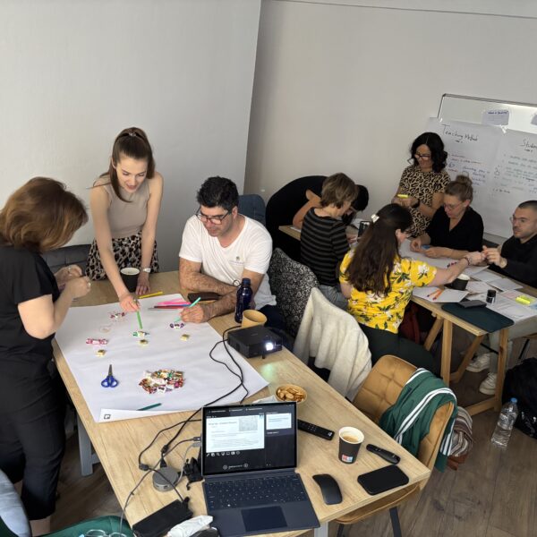 Gruppendiskussion in einem modernen Büro, bei der Teilnehmer an einem Tisch kreativ arbeiten. Auf dem Tisch liegen Bastelmaterialien, Notizen und ein Laptop. Die Atmosphäre ist kollaborativ und inspirierend.
