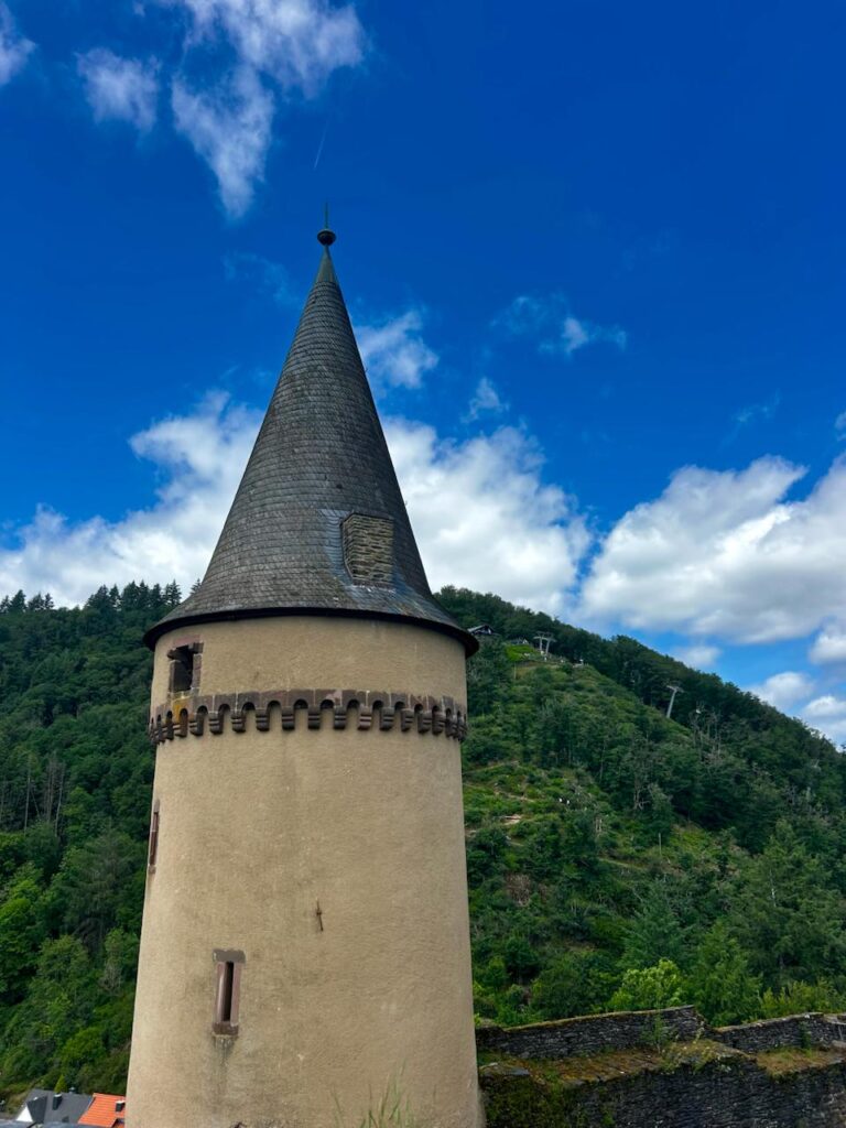 Historischer Turm mit spitzem Dach und Mauerwerk, umgeben von grünen Hügeln und blauem Himmel. Ideal für Architektur- und Naturfotografie.