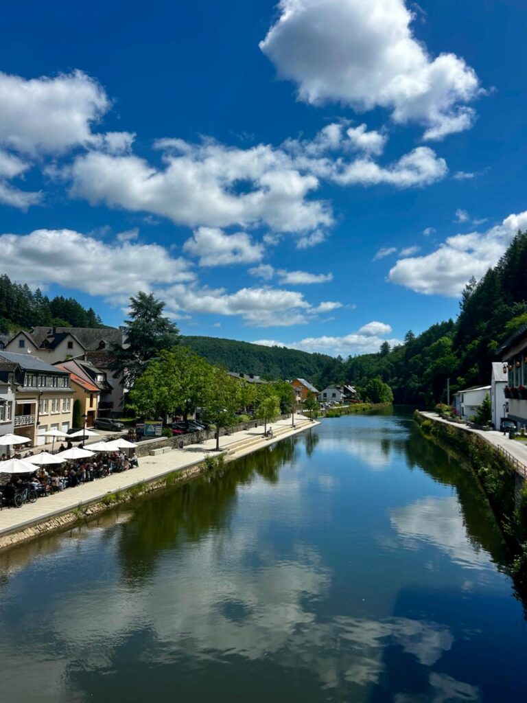 Malerische Flusslandschaft mit reflektierenden Wolken im blauen Himmel, umgeben von charmanten Häusern und grünen Bäumen in einer ruhigen Stadt. Ideal für einen entspannten Spaziergang oder ein gemütliches Café-Erlebnis am Wasser.