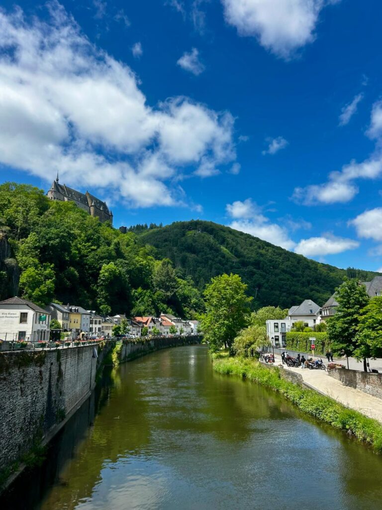 Blick auf die malerische Landschaft mit einem Fluss, umgeben von grünen Hügeln und historischen Gebäuden, darunter eine Burg im Hintergrund und charmante Häuser entlang des Ufers. Strahlend blauer Himmel mit einigen Wolken.