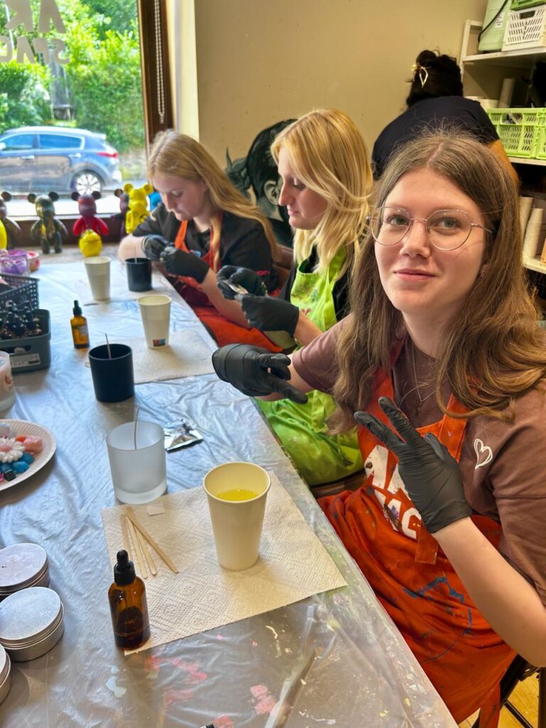 Drei junge Frauen in Schürzen arbeiten an einem kreativen Projekt in einem Atelier. Sie verwenden Handschuhe und konzentrieren sich auf das Mischen von Farben in Bechern. Auf dem Tisch sind verschiedene Malutensilien und dekorative Materialien sichtbar.