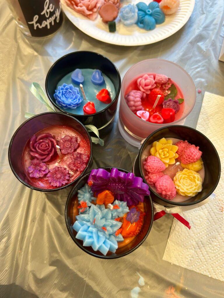 Bunte handgemachte Kerzen mit verschiedenen floralen Designs in schwarzen und transparenten Behältern, auf einem Tisch mit Bastelmaterialien.
