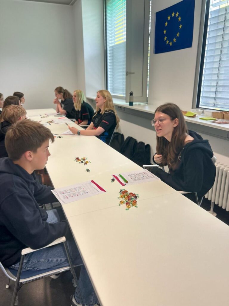 Schülerinnen und Schüler sitzen an einem Tisch in einem Klassenraum, während sie an einem interaktiven Lernprojekt teilnehmen. Auf dem Tisch liegen bunte Spielsteine und Arbeitsblätter. Im Hintergrund hängt die Flagge der Europäischen Union.