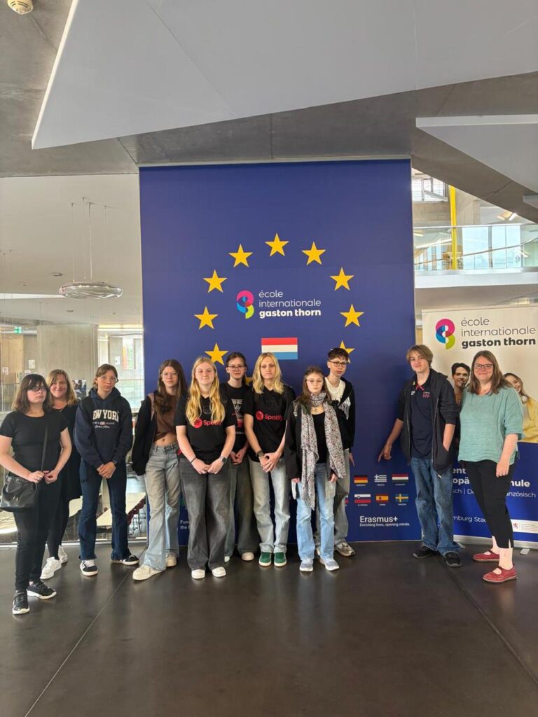 Gruppe von Schülern vor dem Banner der École Internationale Gaston Thorn in Luxemburg, umgeben von europäischen Flaggen und Logos, während einer Schulveranstaltung.