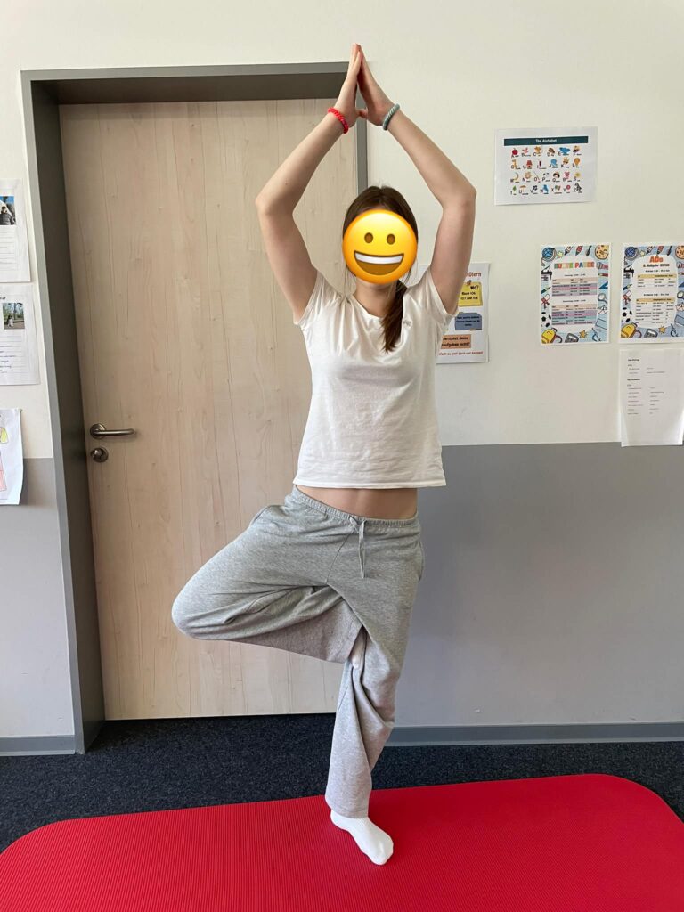 Junge Frau in sportlicher Kleidung, die eine Yoga-Position auf einer roten Matte einnimmt, mit erhobenen Händen und einem Lächeln. Hintergrund zeigt eine Wand mit Informationsblättern und einem Holzportal.