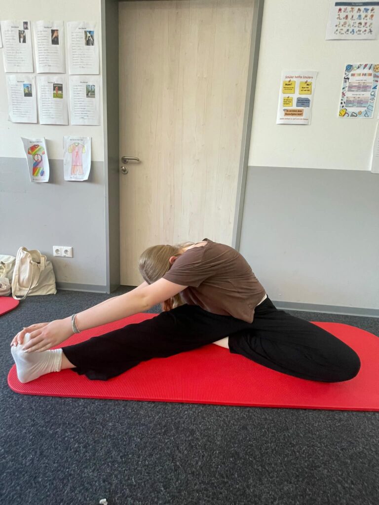 Junge Frau dehnt sich auf einer roten Yogamatte in einem Raum mit Zeichnungen an der Wand. Sie sitzt im Sitzen und streckt ihren Oberkörper nach vorne, während sie einen Fuß greift.