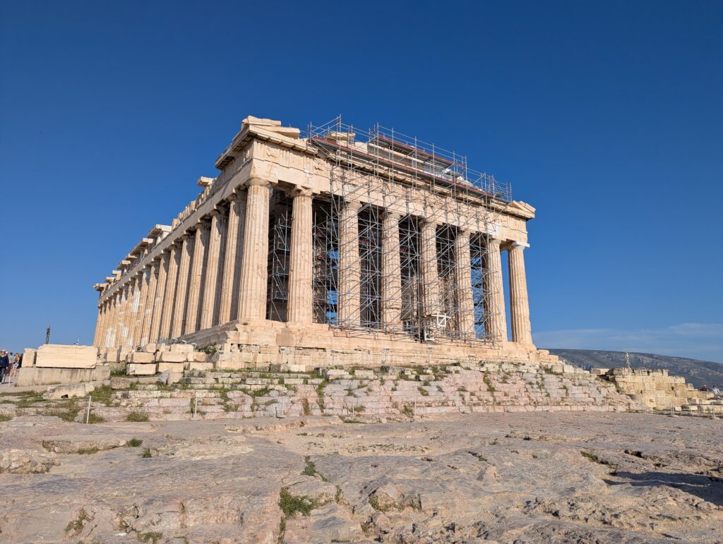 Partenon in Athen, teilweise eingerüstet, unter blauem Himmel, umgeben von antiken Ruinen und Touristen.